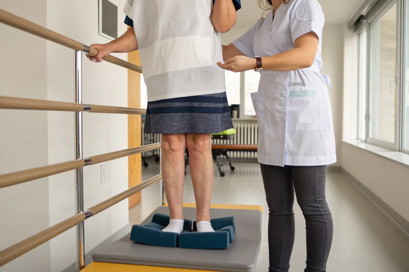 Supported balance exercise on foam pads beside a handrail.